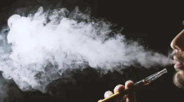 A person blows a cloud of vapor on a dark backdrop after using his vape pen